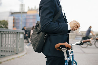 Midsection of businessman holding in-ear headphones while standing with bicycle on bridge in city