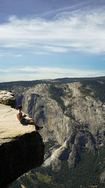 Shirtless man sitting on cliff against cloudy sky