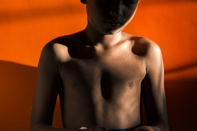 Midsection of shirtless teenage boy standing by orange wall