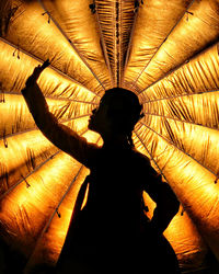 Silhouette woman with hand raised standing against illuminated orange curtain