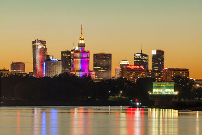 Illuminated buildings by river against sky in city