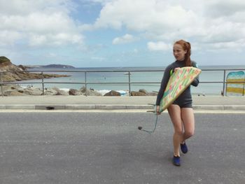 Full length of woman with surfboard walking on road by sea against sky