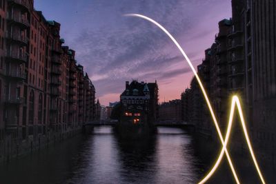 Illuminated light painting over river by buildings against sky in city