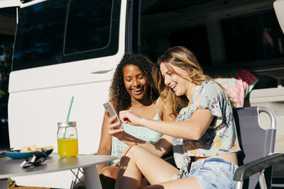 Cheerful women enjoying drink and nachos while sitting out of van in camping using smartphone