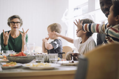 Preschool students gesturing at teacher while sitting at dining table during lunch break