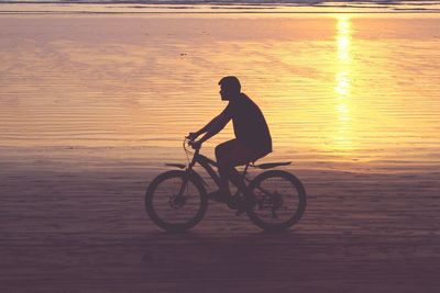 Young man riding bicycle at beach during sunset