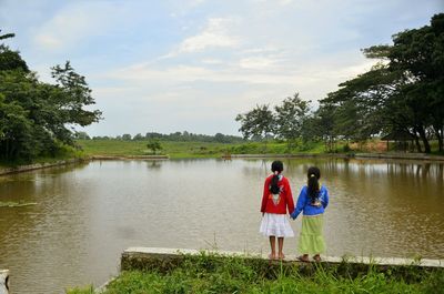 Rear view of siblings standing by river against sky