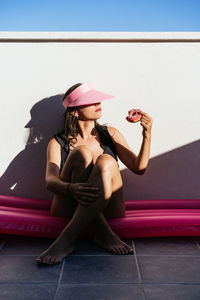Full body of young female in swimsuit and hat sitting on pink inflatable mattress and enjoying delicious donut while chilling on poolside in summer day