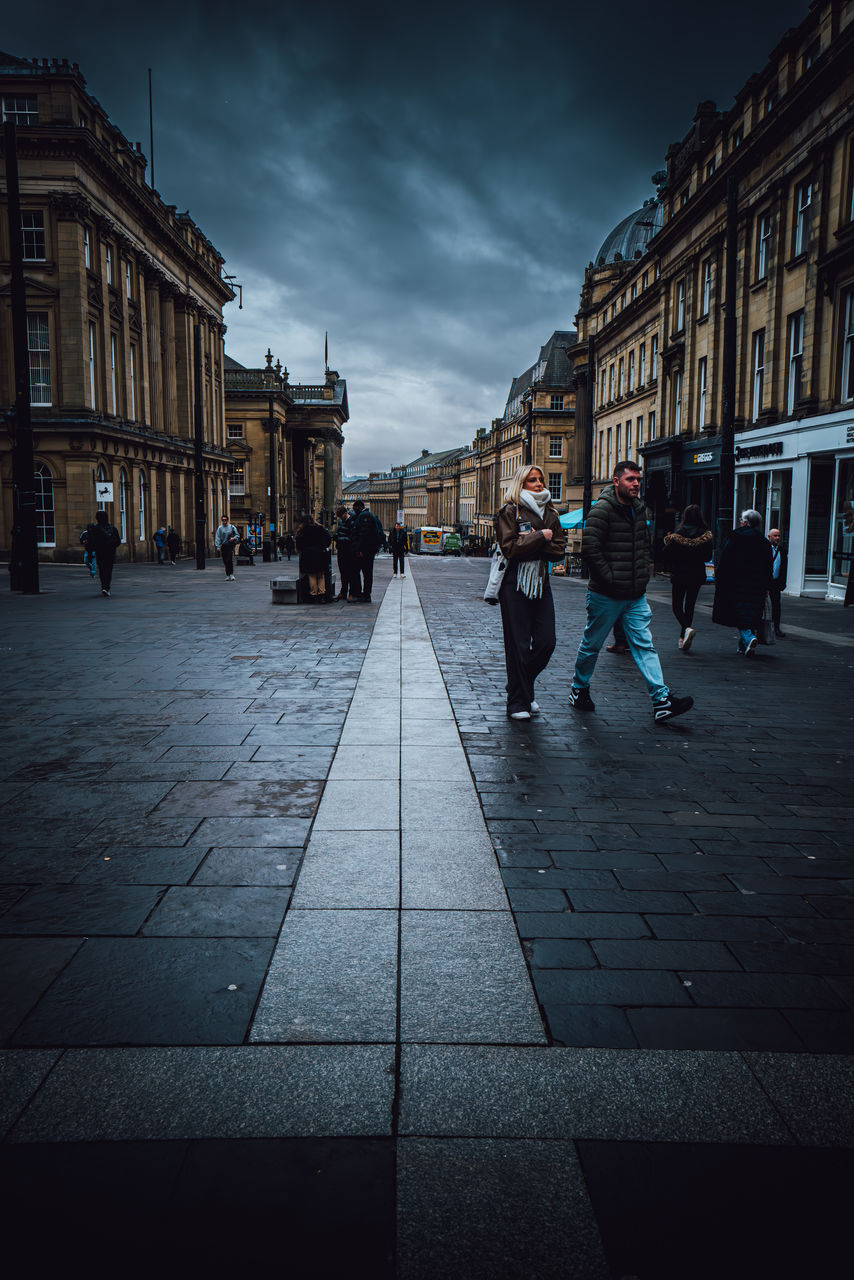 architecture, city, building exterior, darkness, built structure, street, night, group of people, sky, evening, adult, road, walking, light, city life, women, men, urban area, cityscape, building, nature, cloud, travel destinations, full length, city street, cobblestone, footpath, lifestyles, outdoors, reflection, black