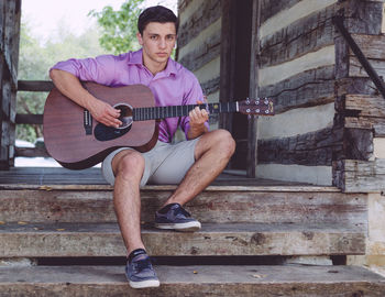 Portrait of man playing acoustic guitar while sitting on steps at log cabin