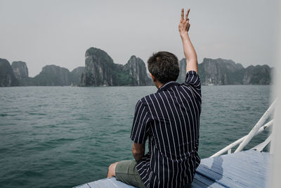 Rear view of man gesturing while sitting in boat at sea against sky