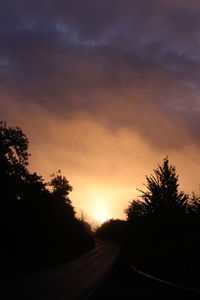 Silhouette trees by road against sky at sunset