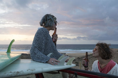 Two surfer girls with sunglasses on their 4x4 car drinking beer sunset