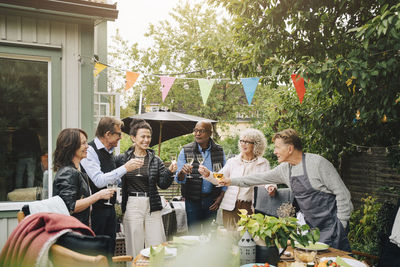 Active senior male and female friends raising toast with drink while standing at back yard during garden dinner party