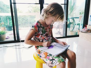 Girl drawing over paper while sitting on stool against window