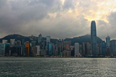 Cityscape by riverside against cloudy sky