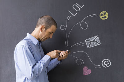 Side view man using mobile phone while standing by blackboard with various icons