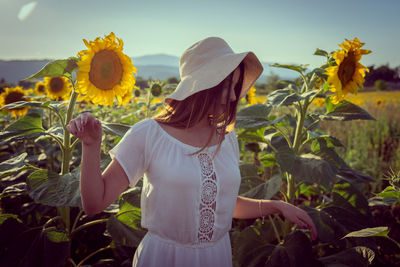 Teenage girl wearing hat standing amidst sunflowers against clear sky