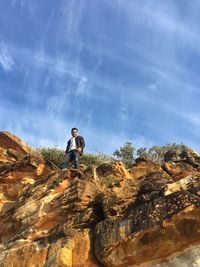 Low angle portrait of young man standing on rock against blue sky during sunny day