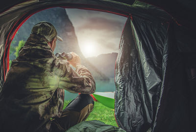 Rear view of man using binoculars while sitting at tent