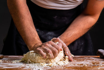 Crop man at wooden table kneading and rolling pile of dough for bread