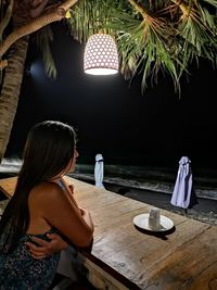 Side view of woman sitting by table at beach during night