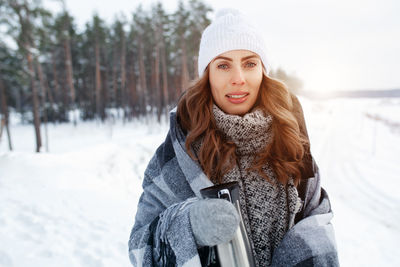 Portrait of confident woman holding insulated drink container while standing in forest during winter