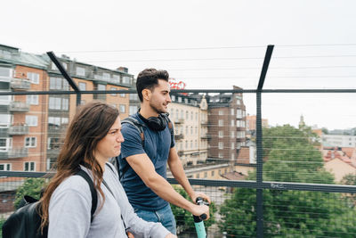 Male and female friends using push scooters on bridge in city against sky