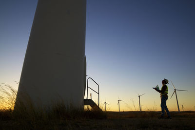 Side view of silhouette engineer looking at windmill while standing on field against clear sky during sunset