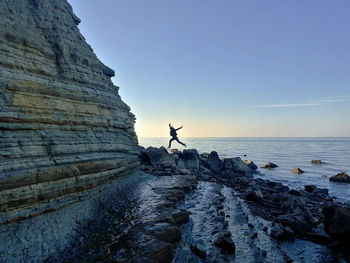 Woman jumping on rocks at beach against sky during sunset