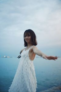 Portrait of smiling young woman standing with arms outstretched
by sea against sky