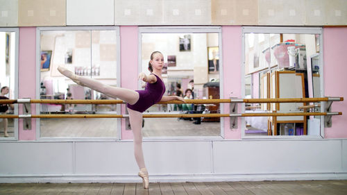 In dancing hall, young ballerina in purple leotard performs developpe attitude on pointe shoes