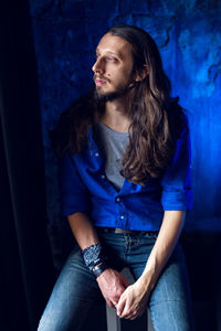 Man with long hair and beard sits at home on a chair by the window in a blue shirt and jeans