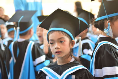 Close-up of girl wearing graduation gown and mortarboard looking away by friends