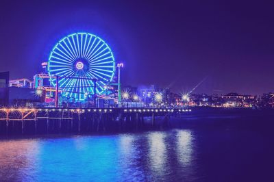 Illuminated ferris wheel by river against sky at night