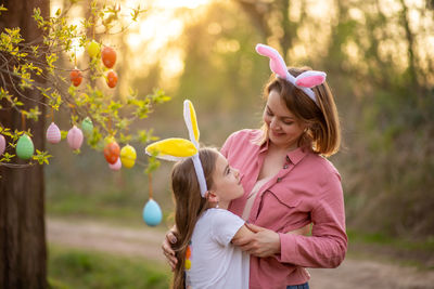 Mother and daughter standing by hanging easter eggs on branch at park