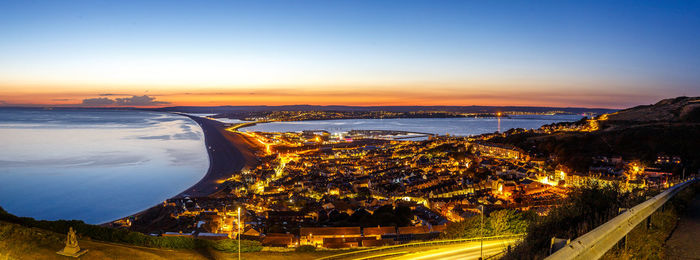 High angle view of illuminated city by sea against sky during sunset