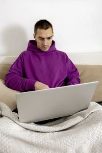 Young caucasian man with violet hoodie sitting on bed and holding laptop computer. 