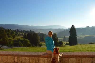 Woman sitting by dog on retaining wall against clear sky