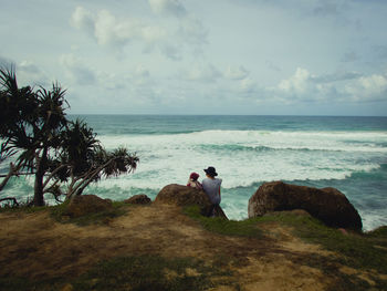 Couple sitting on rock at beach against sky