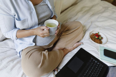 Pregnant businesswoman holding tea cup sitting with laptop on bed