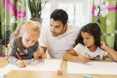 Mid adult teacher assisting female students in drawing at preschool