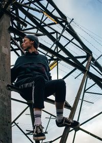 Low angle view of young man sitting on electricity pylon