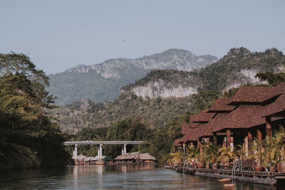 House by river amidst trees and buildings against sky