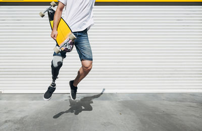Young man with leg prosthesis holding skateboard and jumping