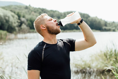 Young man drinking water against lake