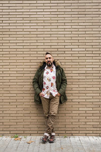 Young bearded man with hands on pocket leaning on wall, looking camera