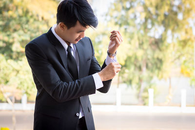 Businessman buttoning shirt while standing against trees