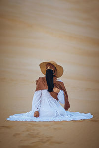 Muslim girl in hijab and niqab wearing a summer hat sitting on the beach sand