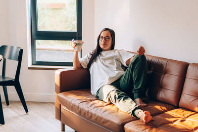 A young brunette woman in glasses and sits on a sofa in the living room holding a cup of drink.